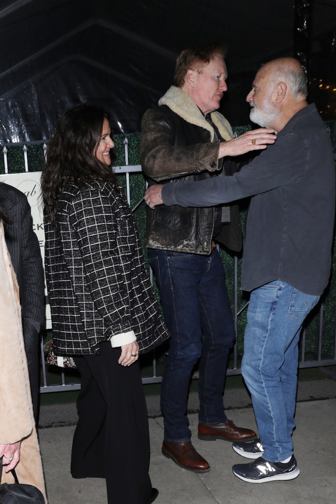 Comedians Rob Reiner and Conan O'Brien talking to each other while a woman wearing a black and white checkered jacket stands to their left, smiling.