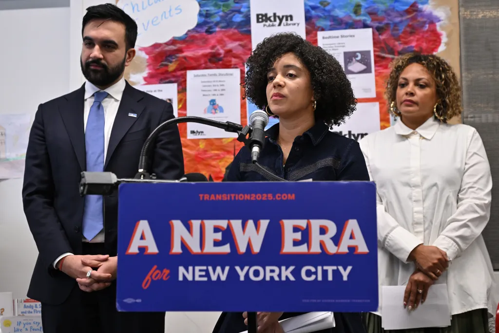 Catherine Da Costa speaking at a podium with “A NEW ERA for NEW YORK CITY” sign, with Zorhan Mamdani and Jahmila Edwards standing beside her.