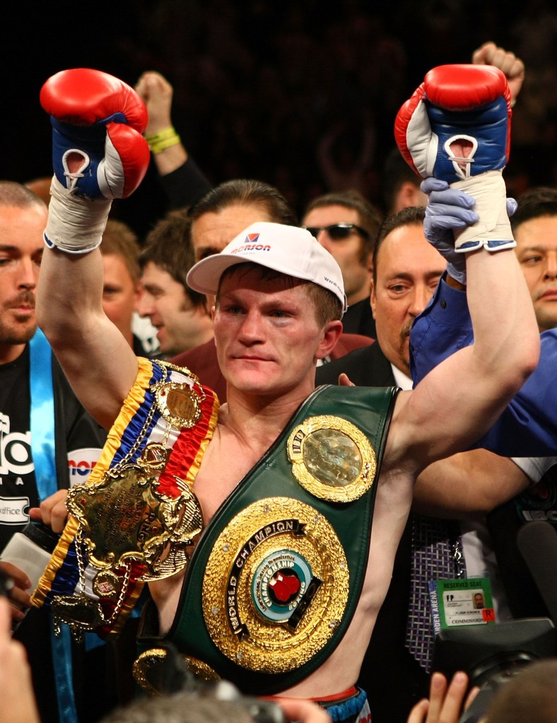 Ricky Hatton celebrates his light-welterweight TKO victory with two championship belts and his arms raised.