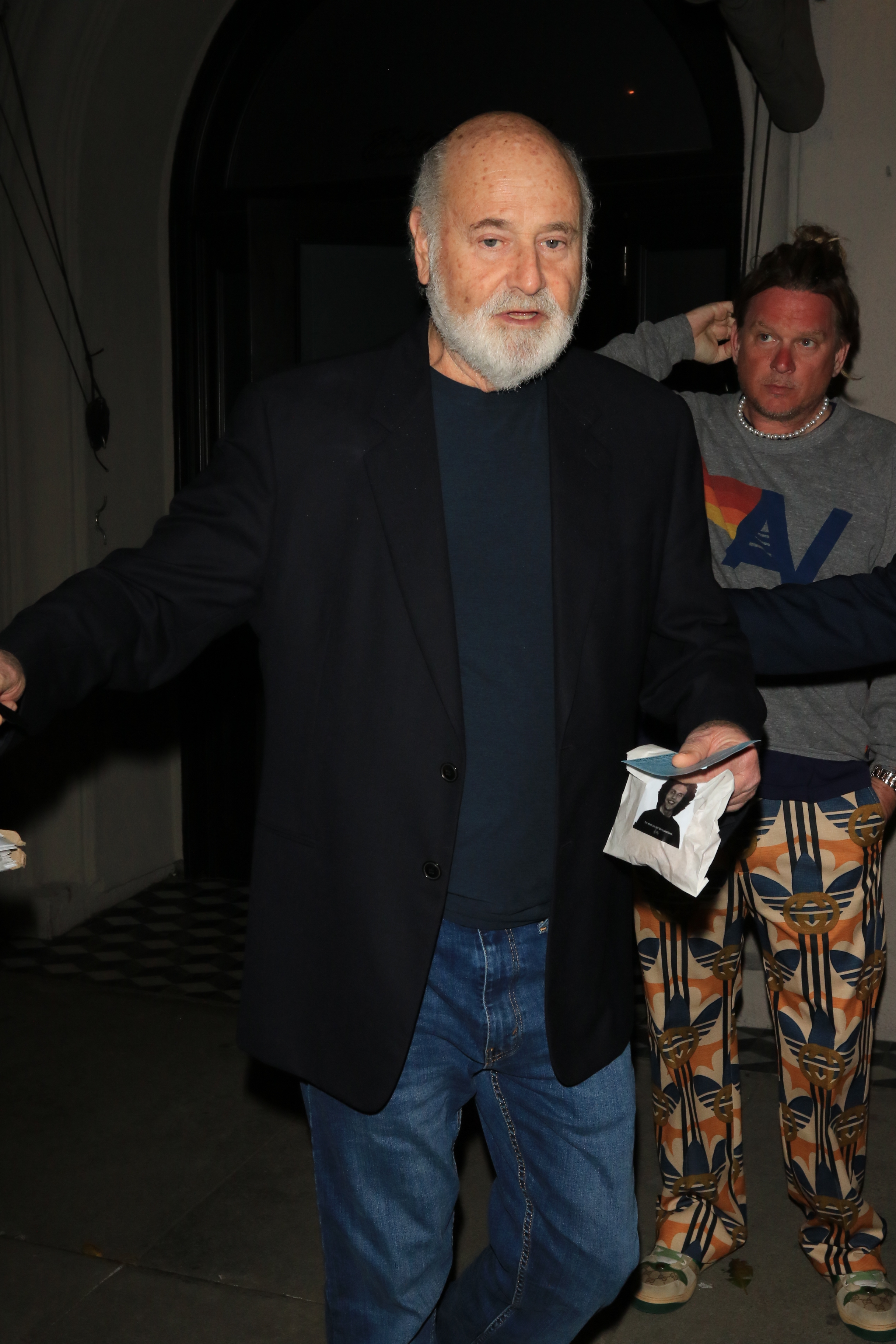 Rob Reiner, wearing a black jacket and jeans, stands next to a fan and holds a piece of paper with an image of himself.