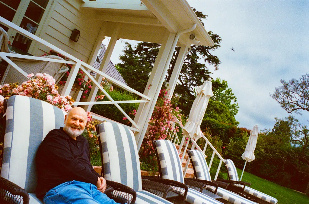 Rob Reiner smiling while seated in an outdoor lounge chair with a light blue and white striped cushion.