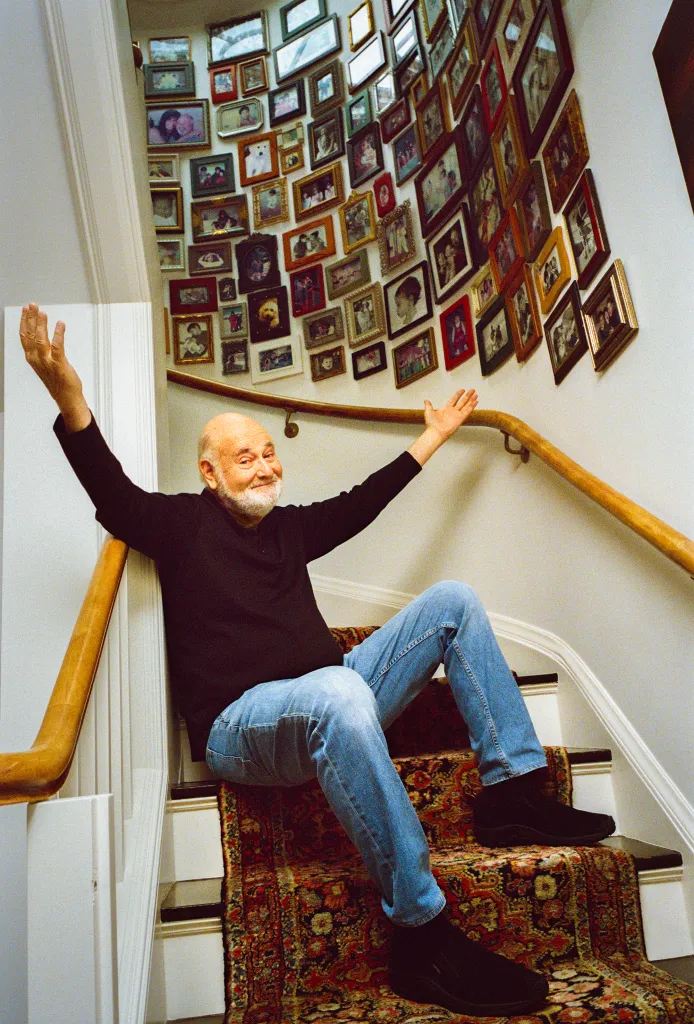Rob Reiner sitting on a carpeted staircase with many framed photos on the wall.
