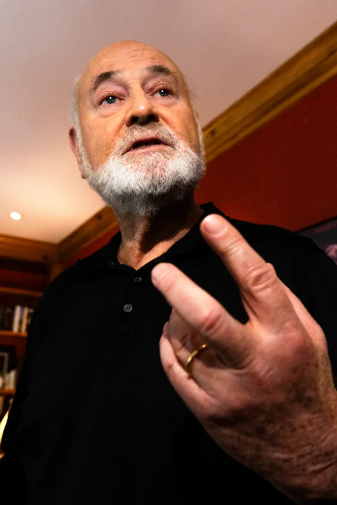 Rob Reiner, bald with a white beard, looks up and points two fingers.