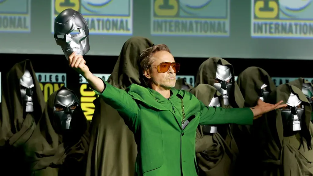 Robert Downey Jr. stands on stage at Comic-Con in a Victor Doom costume, hold his mask toward the sky in a triumphant gesture