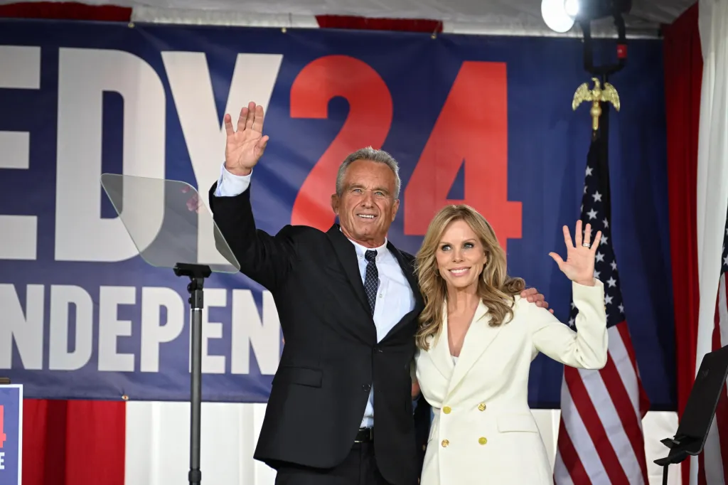 Robert F. Kennedy Jr. in a black suit and black tie, and Cheryl Hines, in a white pant suit, wave to supporters after he announced his independent candidacy for the 2024 presidential race.