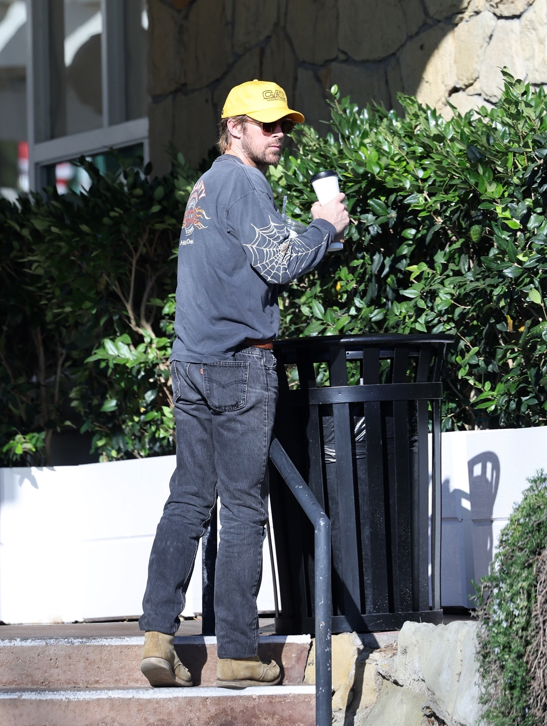 Ryan Gosling wearing a long-sleeved gray shirt with spiderwebs, a yellow baseball cap, and jeans, throwing trash in a bin.