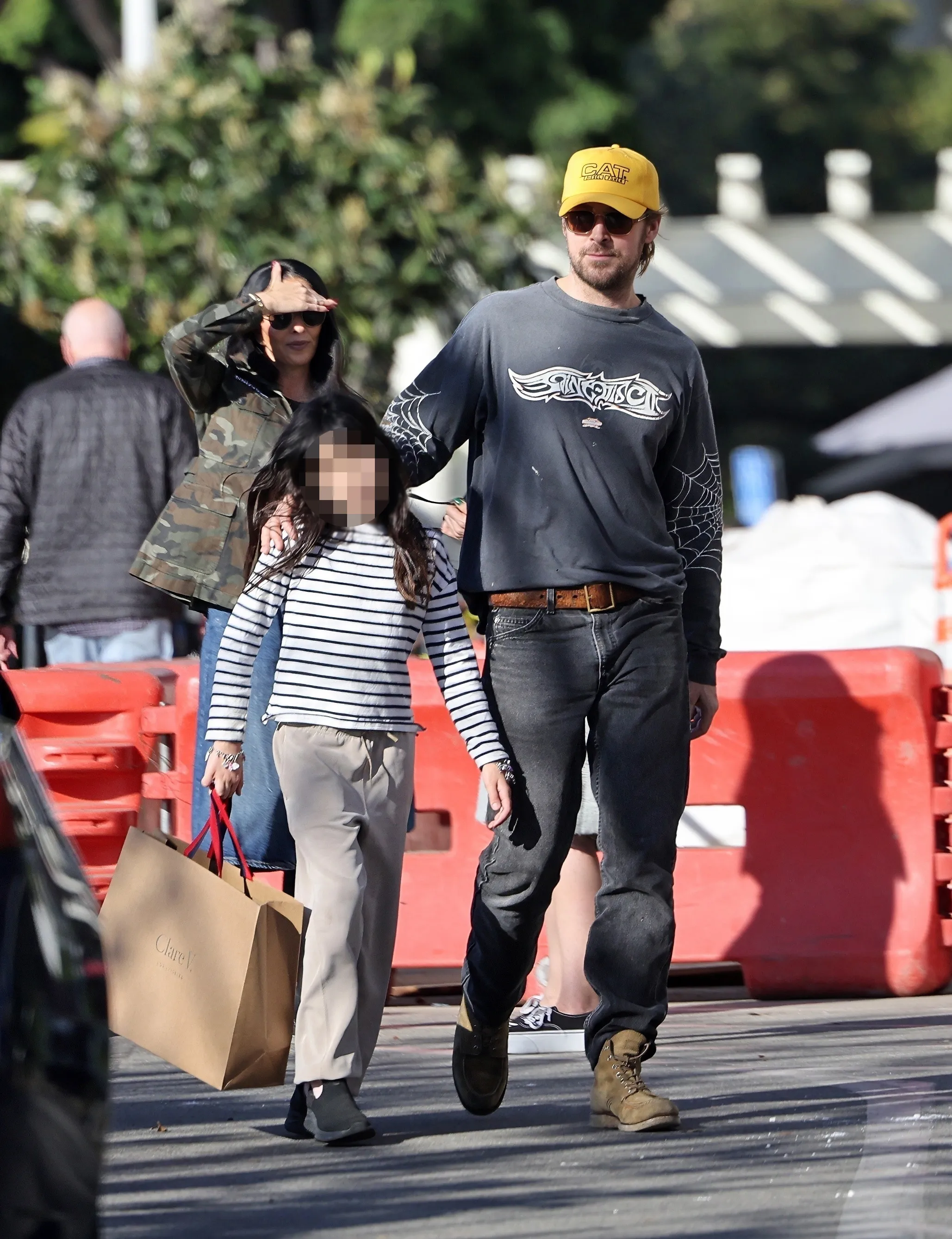 Ryan Gosling gently leads his daughter Amada, who carries a shopping bag, while Eva Mendes walks behind them shielding her eyes from the sun.