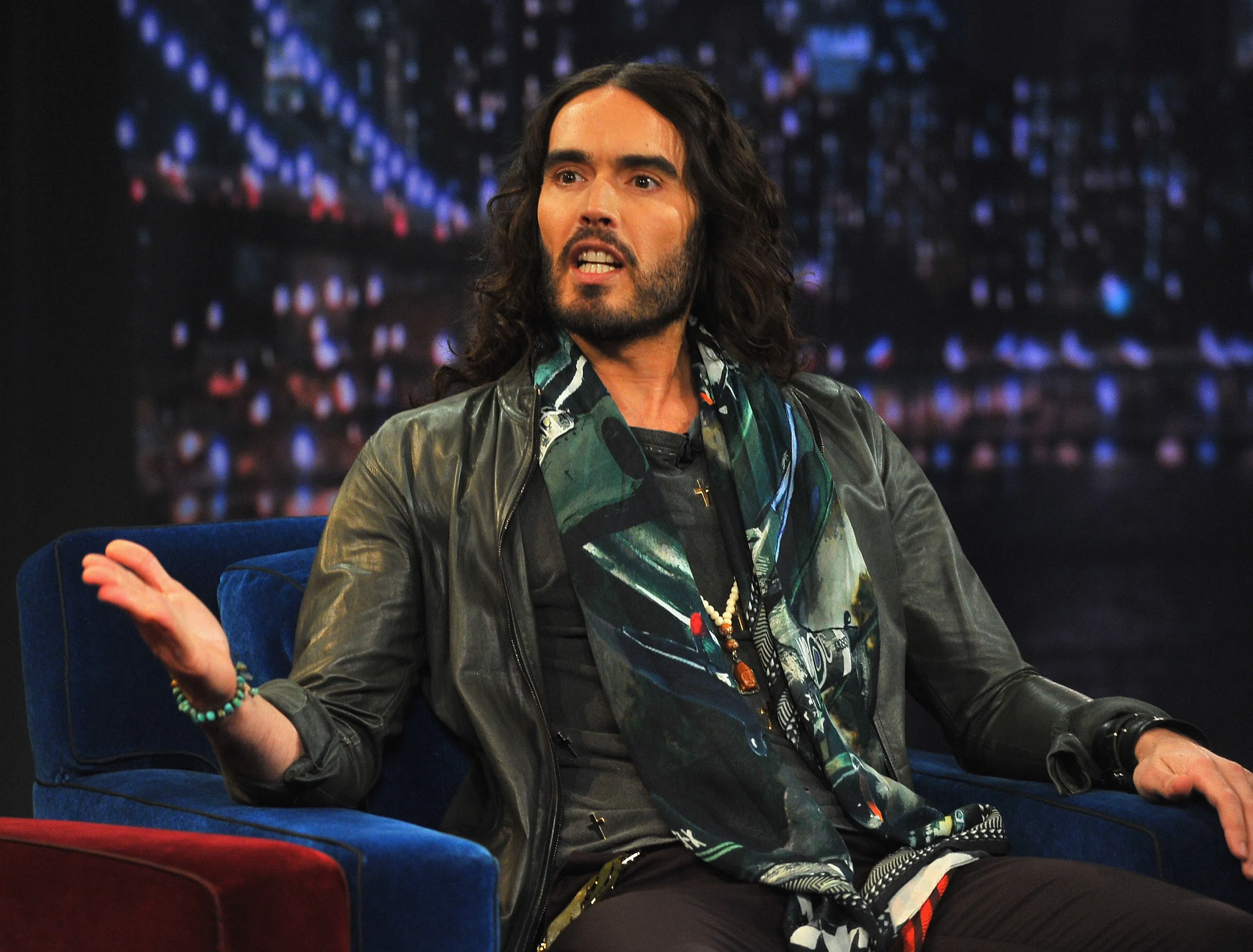 Russell Brand sitting on a blue couch, talking.