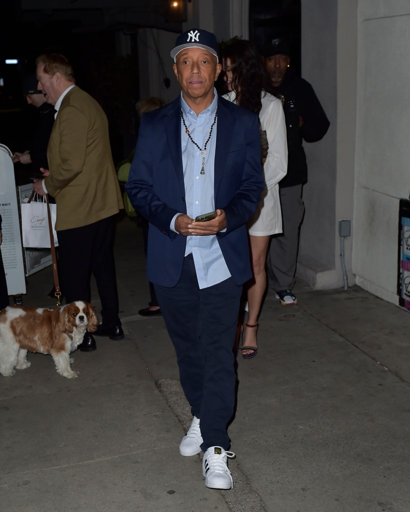 Russell Simmons wearing a Yankee baseball cap, blue jacket, and beaded necklace, holding a phone, walking on a sidewalk.