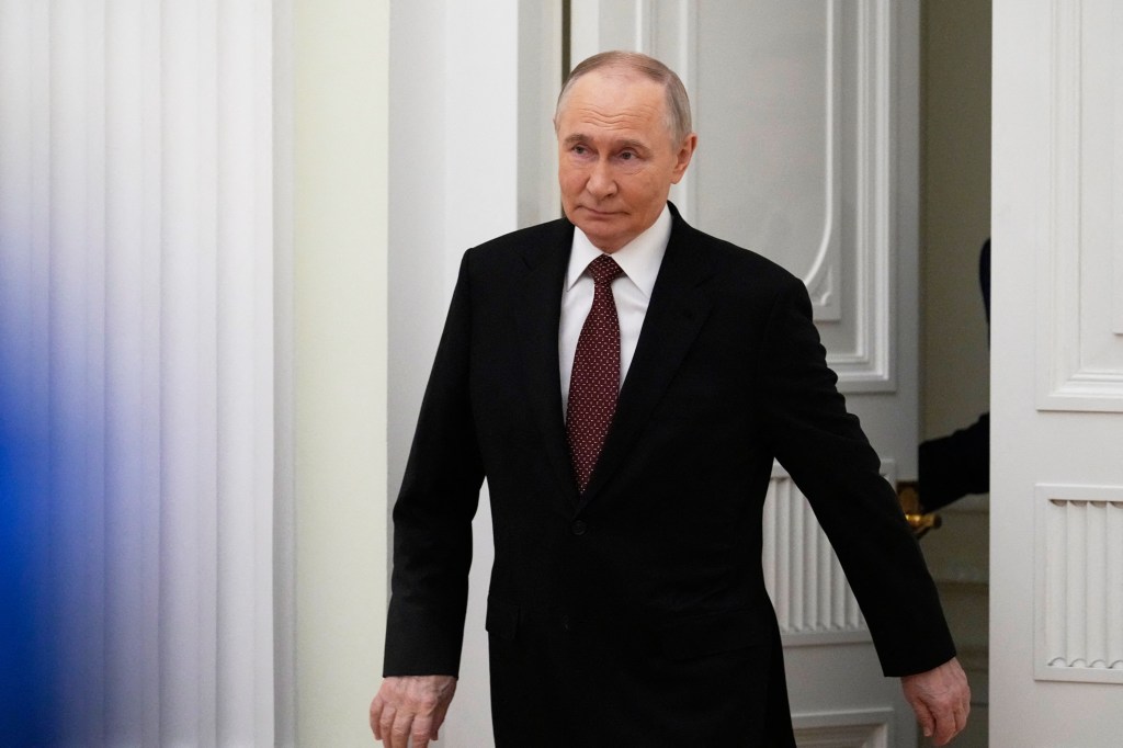Russian President Vladimir Putin wearing a dark suit and patterned red tie, walking into a room.