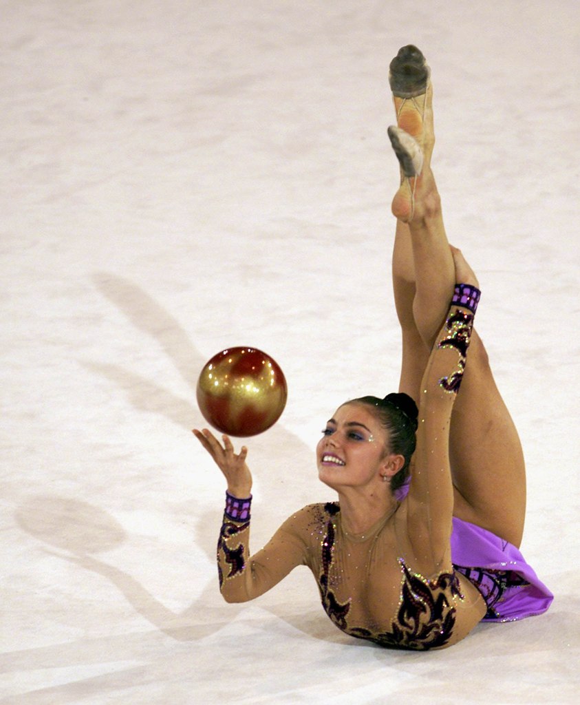 Alina Kabaeva performing a rhythmic gymnastics routine with a ball, arched on her back with legs raised, wearing a sparkly leotard.