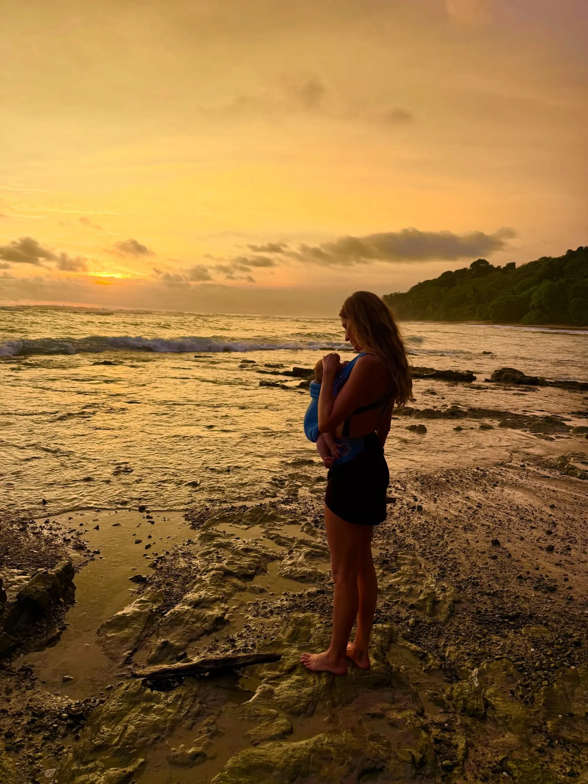 Gisele Bündchen holding a baby in a carrier on a rocky beach at sunset.