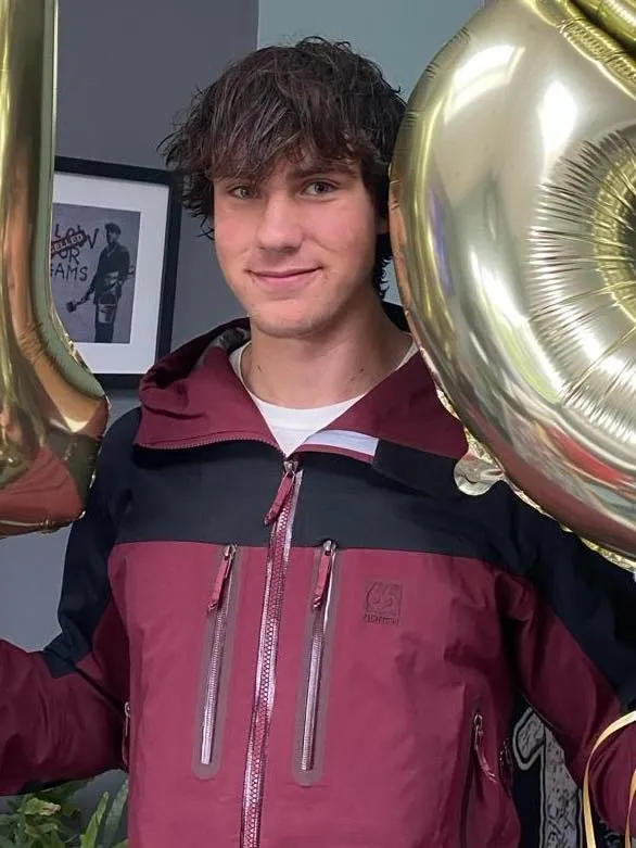 George Smyth, a young man with brown curly hair, smiling and wearing a black and maroon jacket, holding gold balloons.