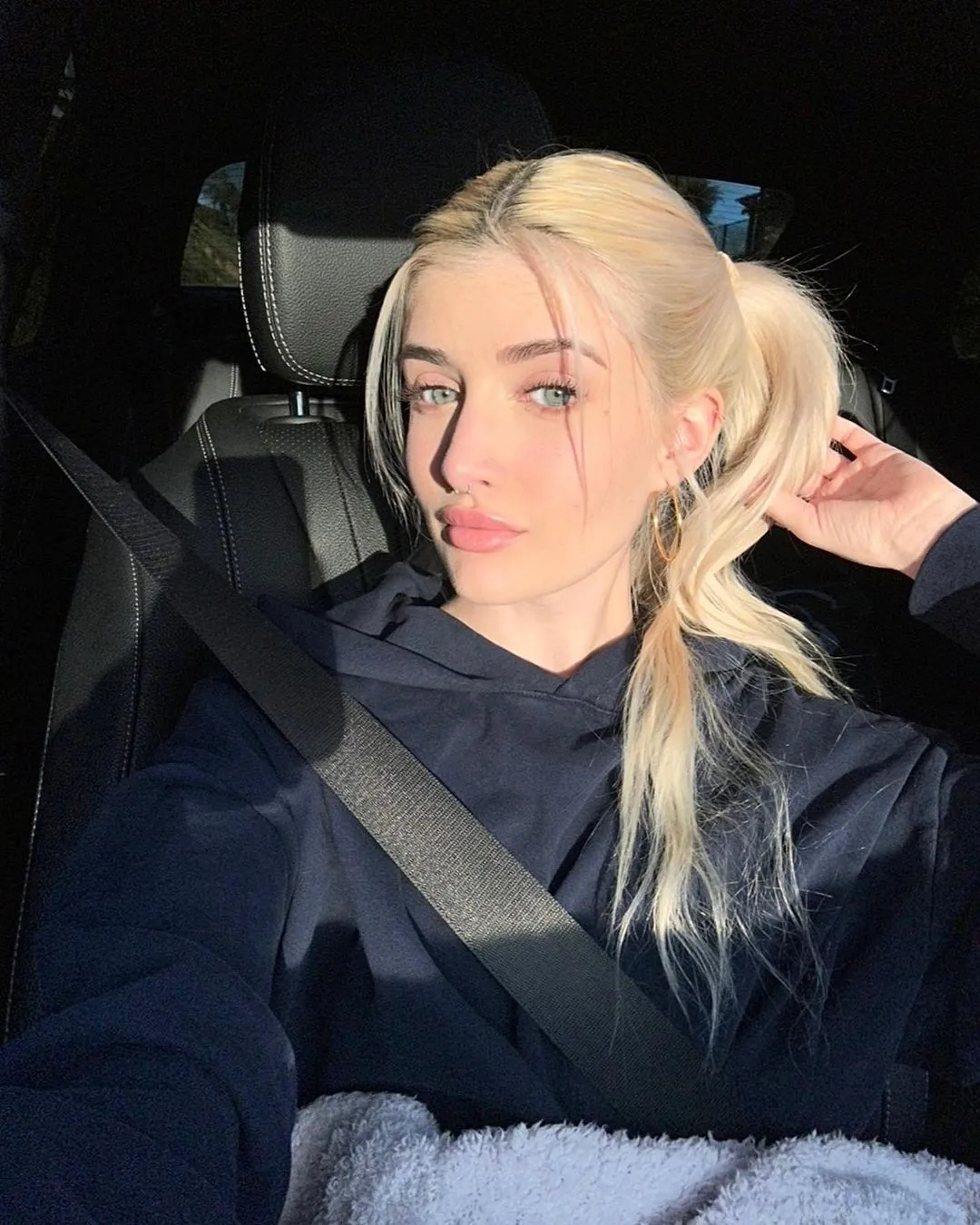 Sami Sheen in a car, wearing a seatbelt and a dark hoodie, with blonde hair tied in a ponytail.