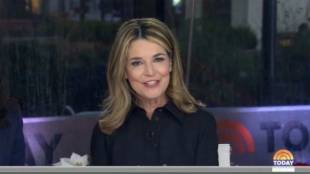 Savannah Guthrie with long blonde hair, wearing a black shirt, speaks during a broadcast.
