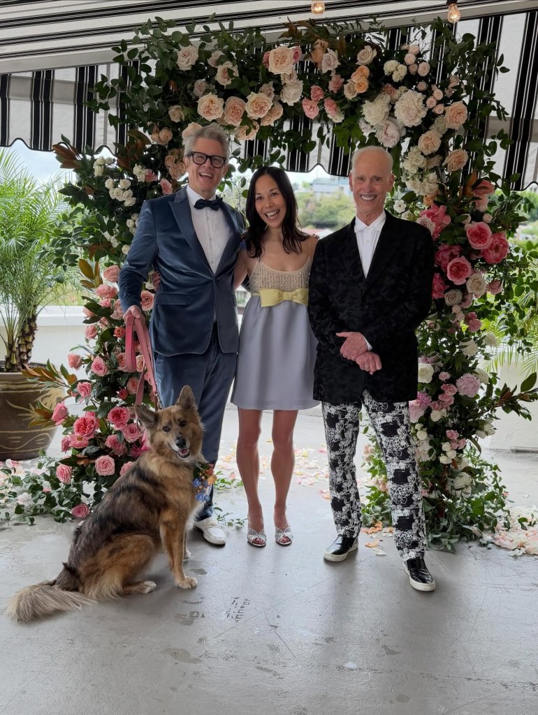Johnny Knoxville, Emily Ting, and John Waters stand with a dog under a floral arch.