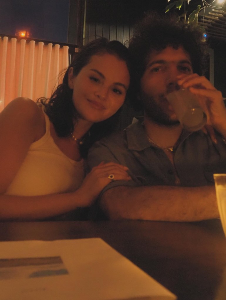 Selena Gomez sitting next to a man at a table, smiling at the camera, with her hand on the man's arm.