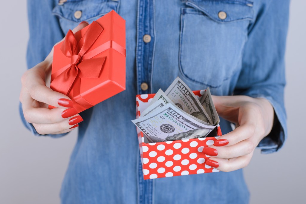 Person holding a red gift box with a red bow and a red and white polka dot box filled with US 100 dollar bills.