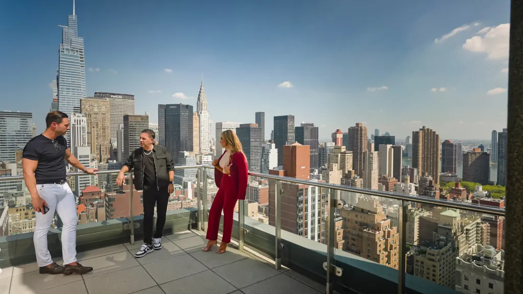 Eleonora Srugo, in a red suit, with two men on a balcony overlooking the New York City skyline.