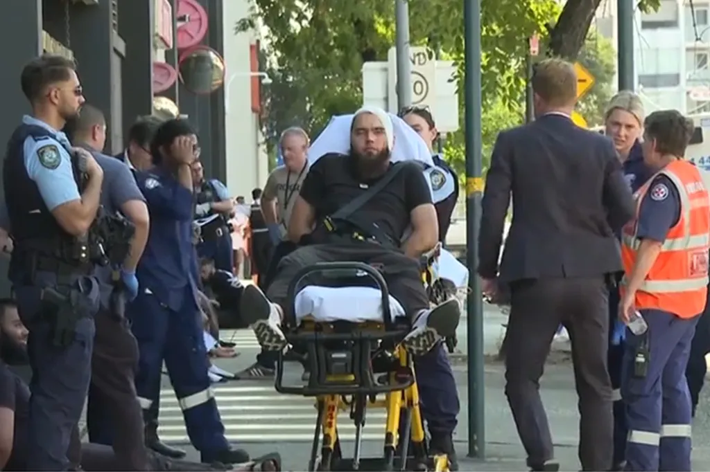 A man with a bandaged head being transported on a stretcher by paramedics, surrounded by police officers.