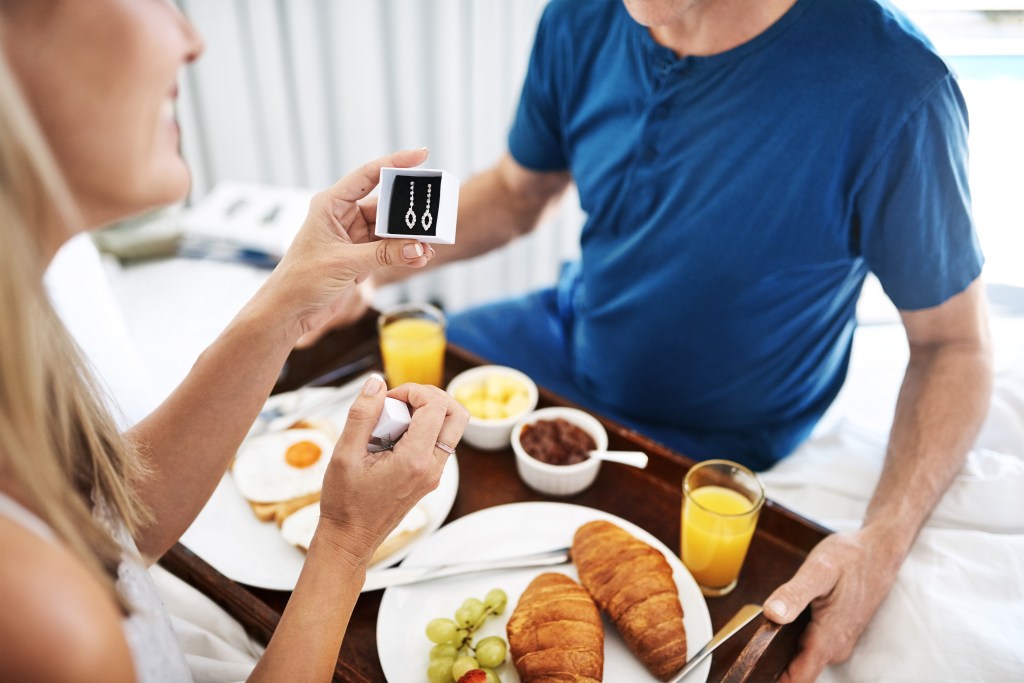 A woman in bed holding up a pair of diamond earrings given to her by a man, with breakfast on a tray between them.
