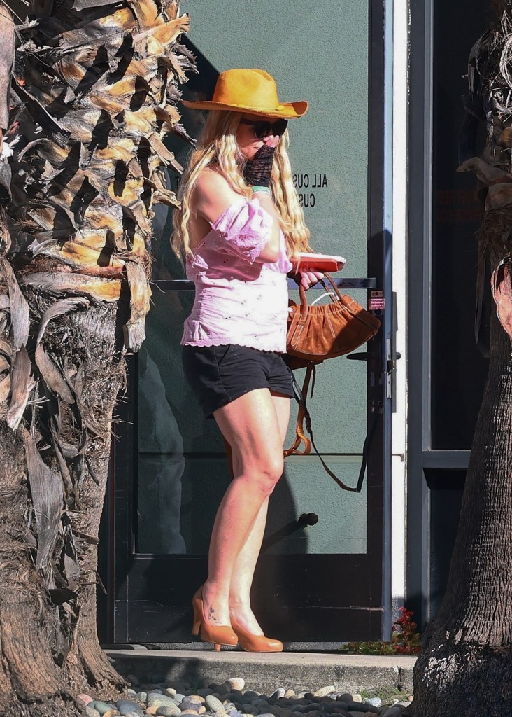 Britney Spears shielding her face with her hand, wearing a wide-brim hat, sunglasses, a pink off-shoulder top, black shorts, and heels.