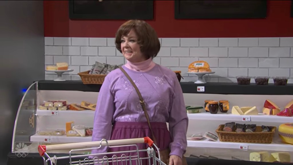 Melissa McCarthy in a lavender shirt and purple skirt, standing next to a grocery cart in a deli aisle.