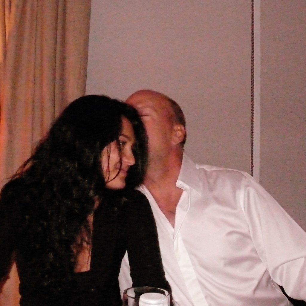 Emma Heming smiling as Bruce Willis kisses her head.