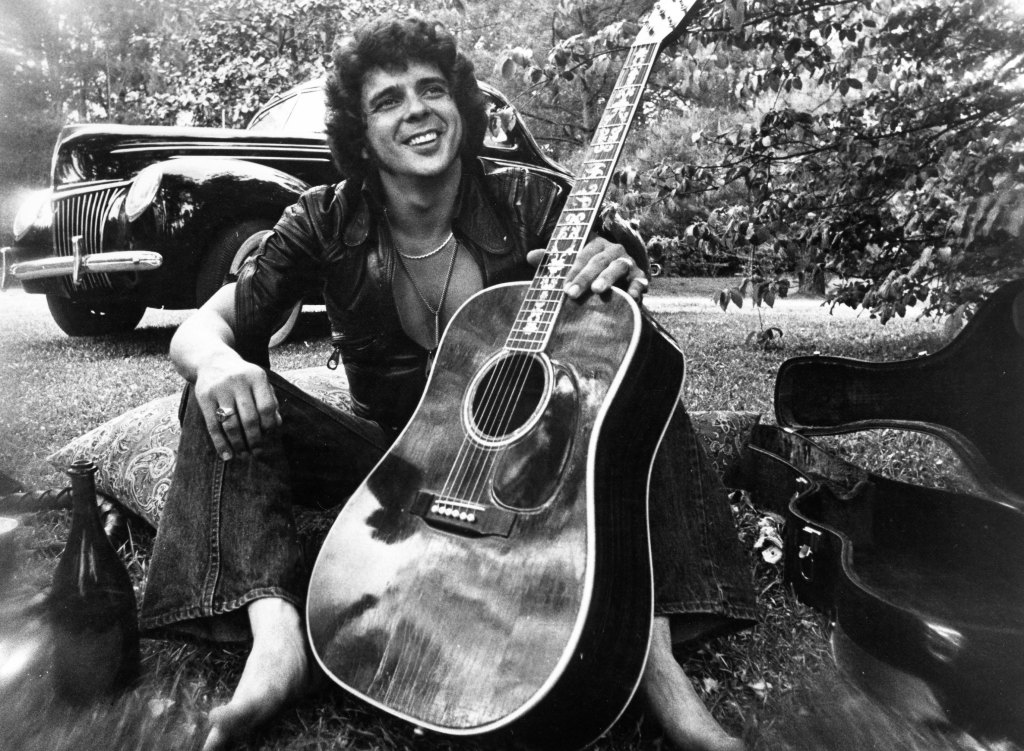 Songwriter and guitarist Bobby Whitlock poses for a portrait in front of an old car next to a bottle of Dom Perignon champagne holding an acoustic guitar in circa 1975.