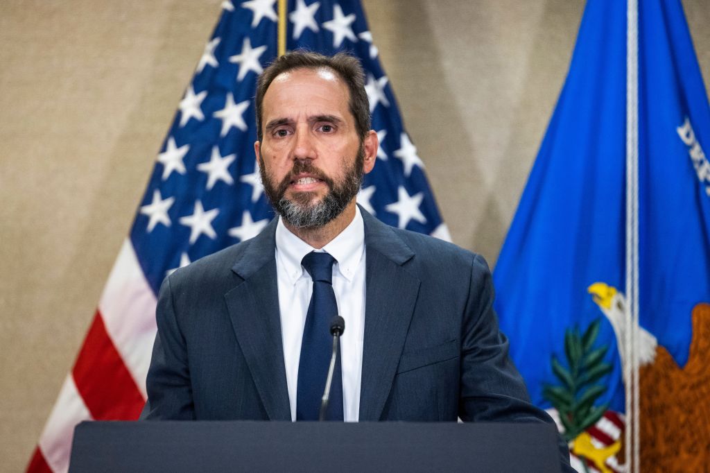 Special Counsel Jack Smith speaking at a podium with American and Department of Justice flags behind him.