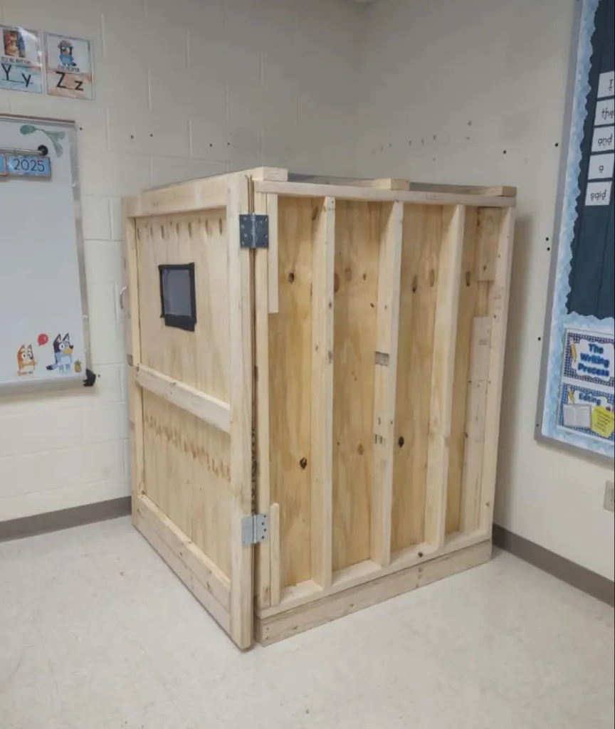 A wooden box, resembling a crate with a small, darkened window, stands in a classroom.