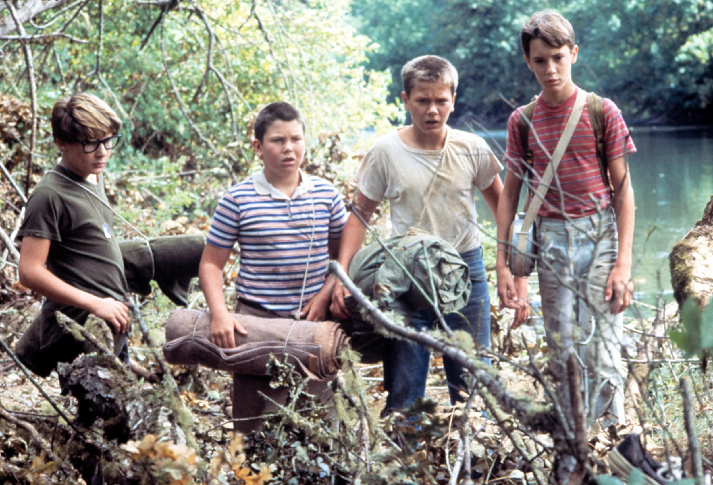 Four young boys, including Corey Feldman and Jerry O'Connell, standing in a wooded area with a river behind them.