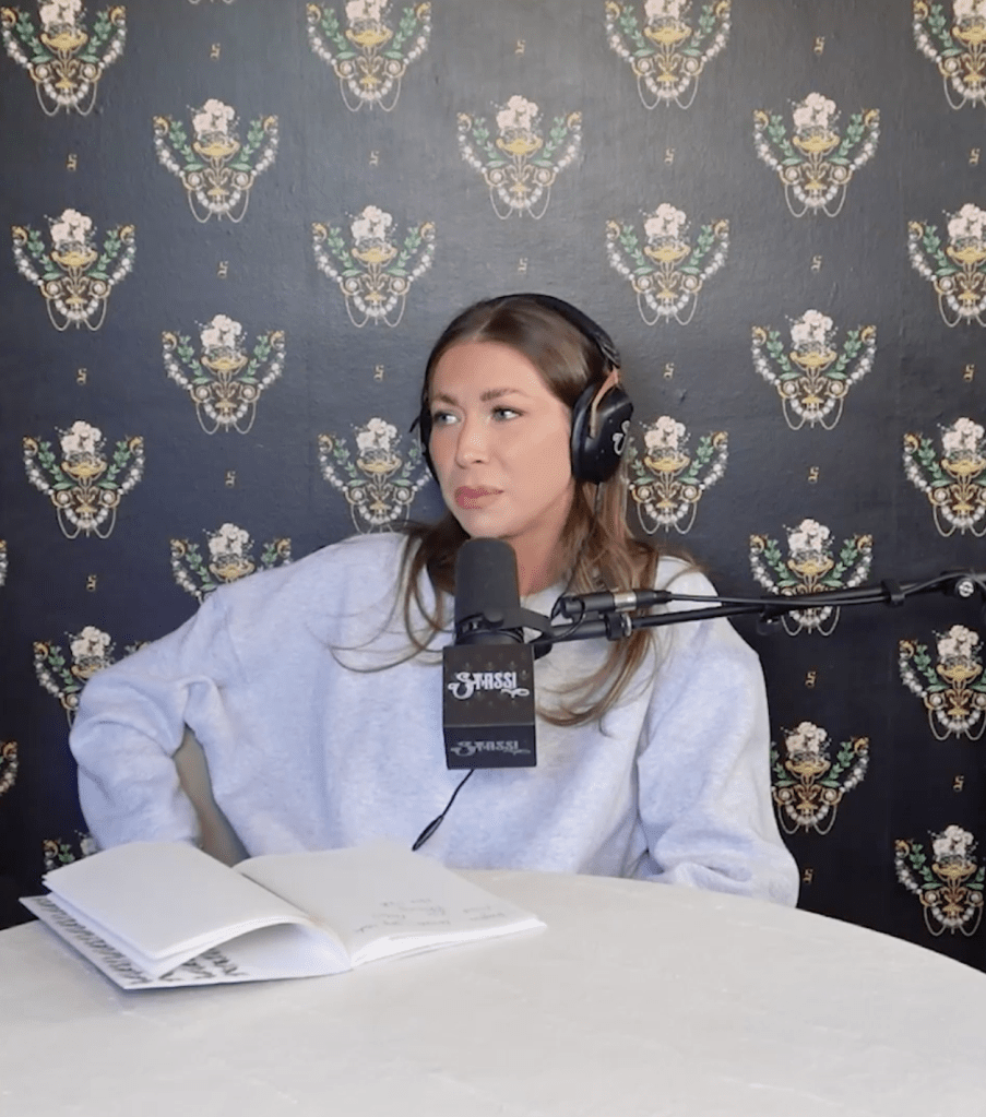 Stassi Schroeder wearing headphones and a grey sweatshirt, speaking into a microphone.