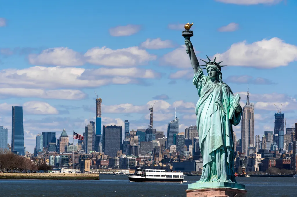 The Statue of Liberty in the foreground with the New York City skyline and the Hudson River in the background.