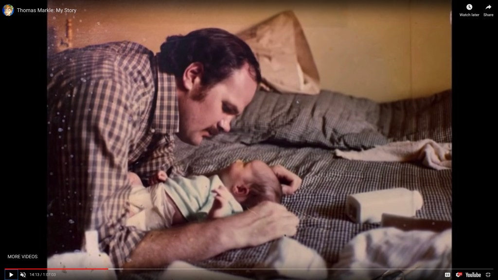 Thomas Markle, in a checked shirt, leaning over baby Meghan Markle laying on a patterned bedspread.