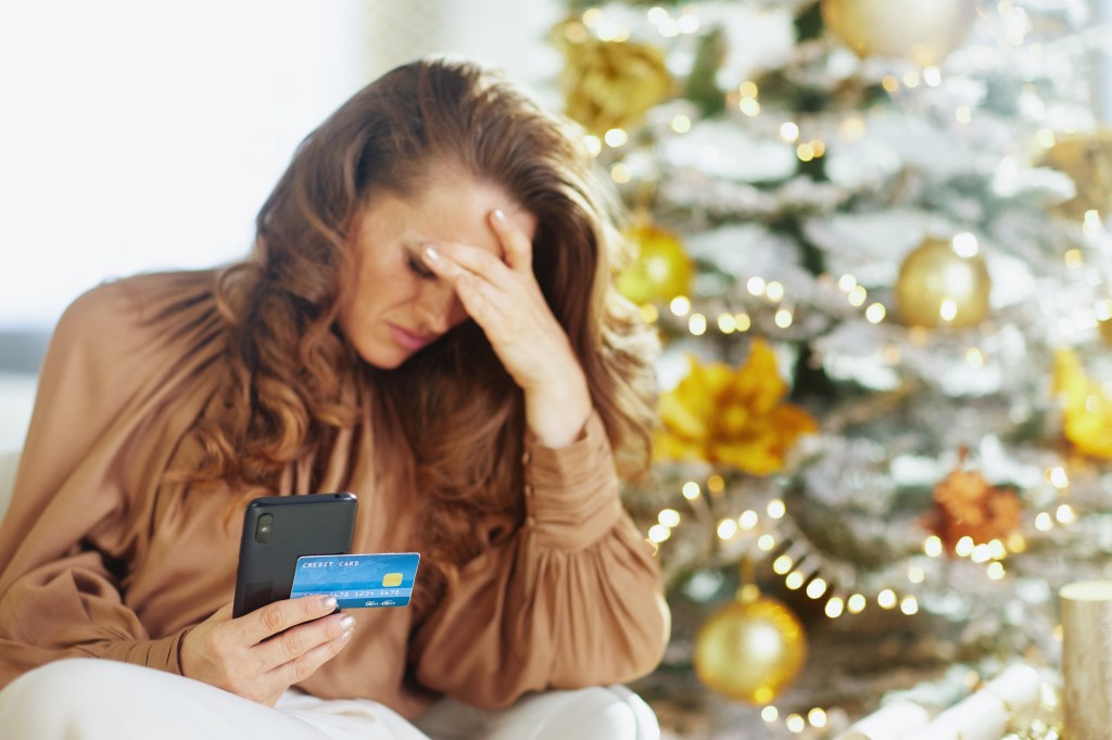 A stressed woman holding a smartphone and credit card in front of a Christmas tree, representing holiday financial anxiety.