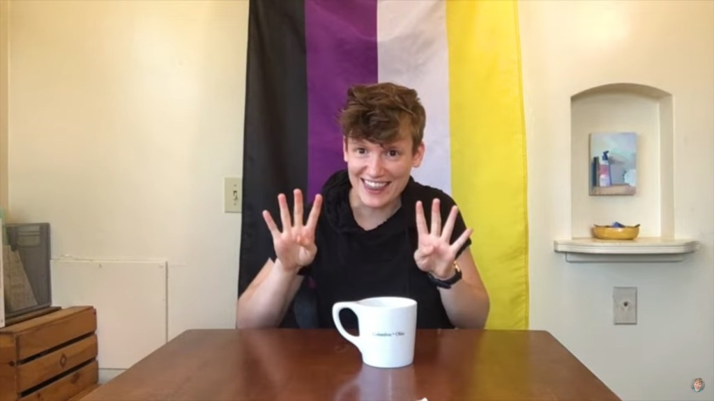 A person in a black shirt gestures with both hands in front of a non-binary flag.