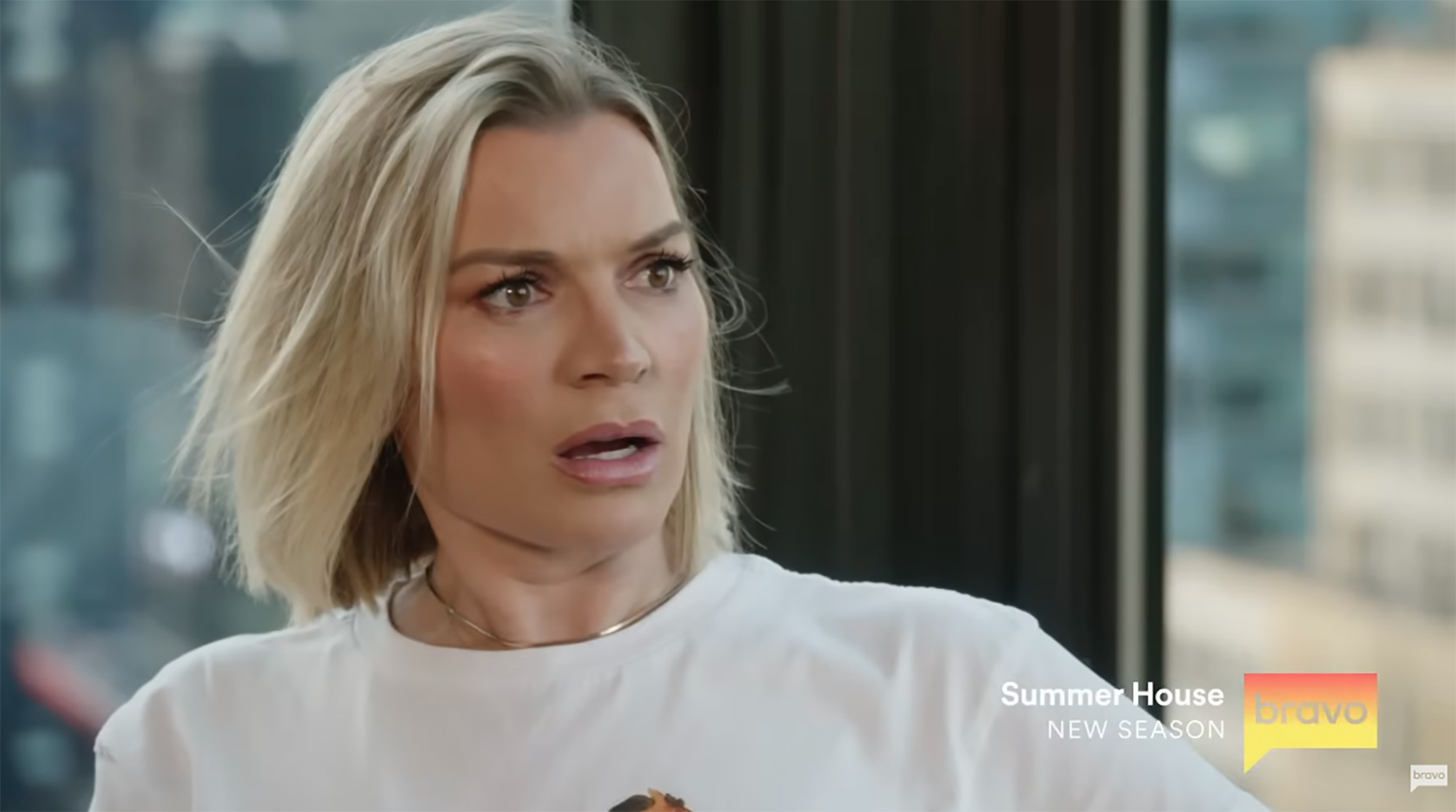 A woman with blonde hair, wearing a white shirt and gold necklace, with an astonished expression.