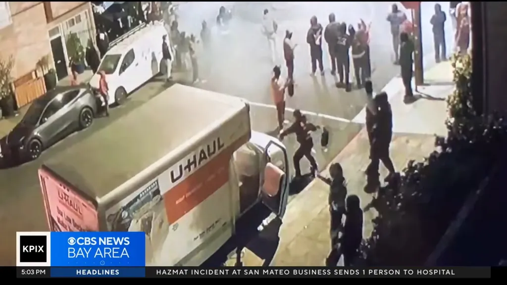 Security camera footage of a large group of people gathered in a street, with a U-Haul truck and other cars parked nearby.
