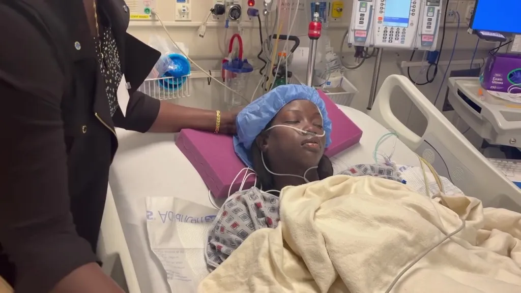 Anok Yai in a hospital bed with a nasal cannula and a surgical cap, with a person's arm resting on her head.