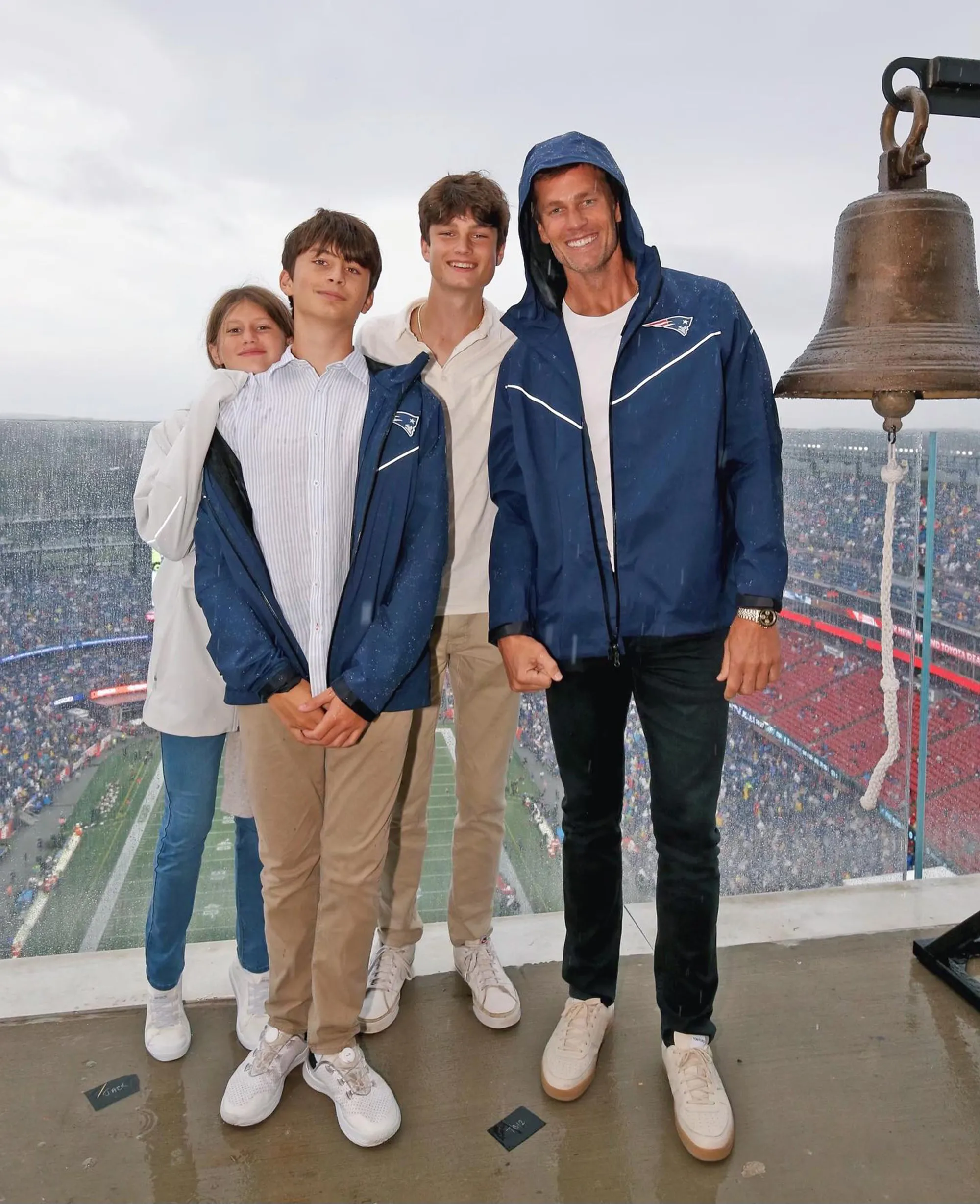 Tom Brady stands with his three children under a bell at a football stadium.