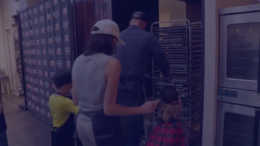 Prince Harry and Meghan Markle with their children, moving a cart of baked goods.