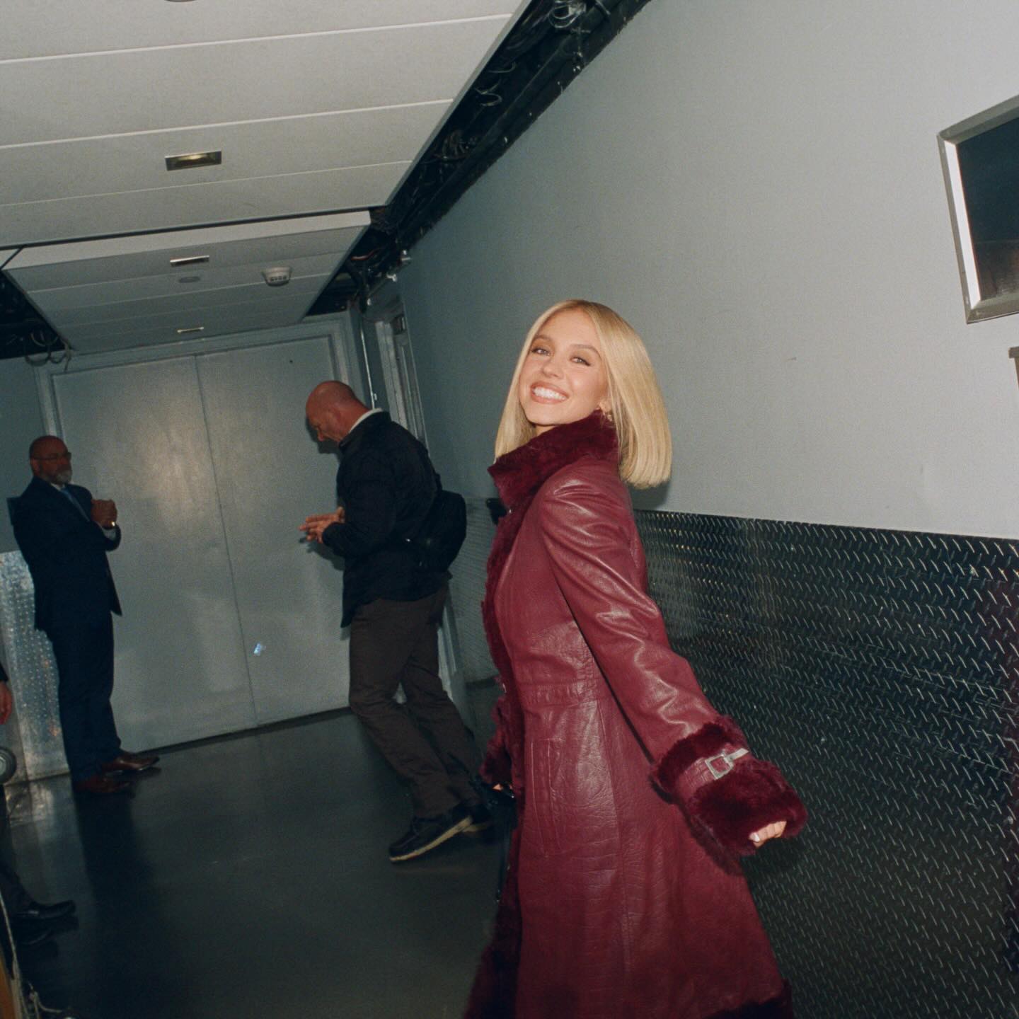 Sydney Sweeney in a long red leather coat with fur trim, smiling over her shoulder in a backstage hallway.