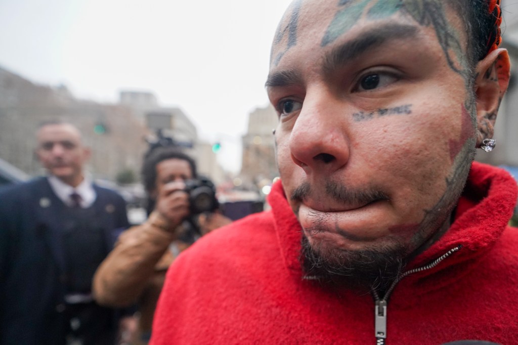 Close-up of Takashi 6ix9ine with multiple facial tattoos, wearing a red fuzzy hoodie, outside 60 Foley Square Courthouse.