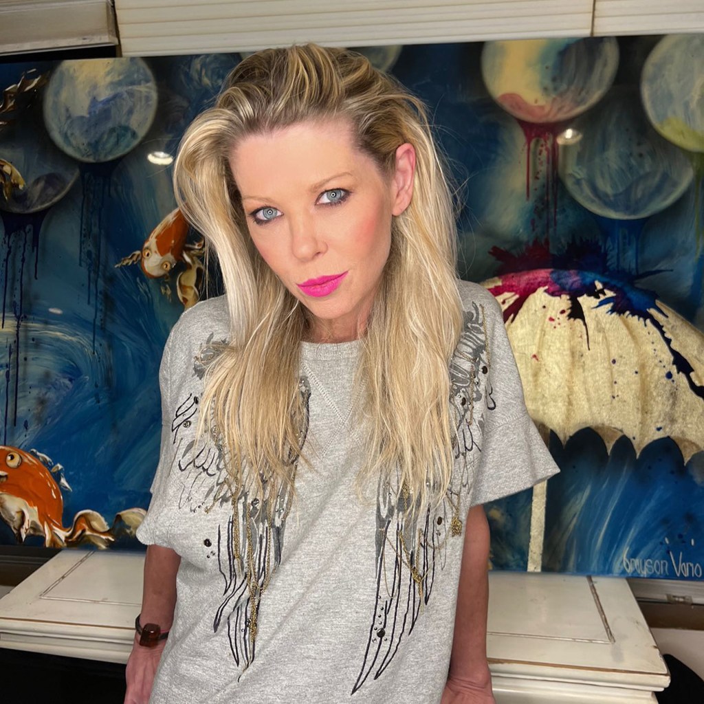 Tara Reid with long blonde hair and bright pink lipstick wearing a gray shirt with black and gold wing designs.