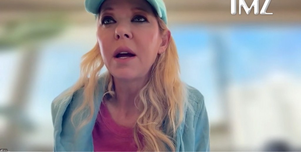 Actress Tara Reid, wearing a turquoise hat and light blue shirt, looks upwards with her mouth slightly open, appearing to speak.