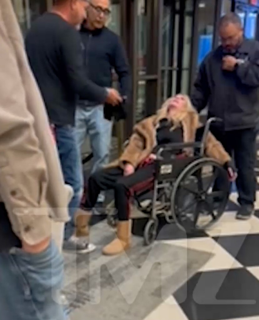 Tara Reid slumped in a wheelchair, being attended to by three men.