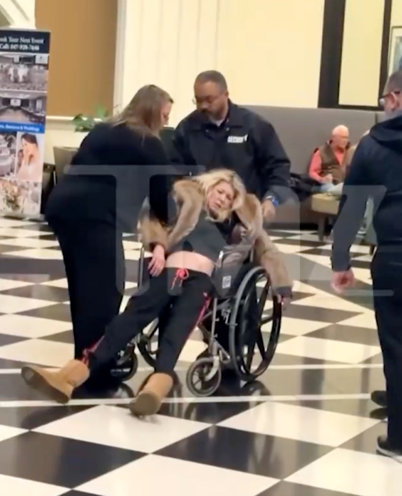 Tara Reid semi-conscious in a wheelchair, assisted by a man in a