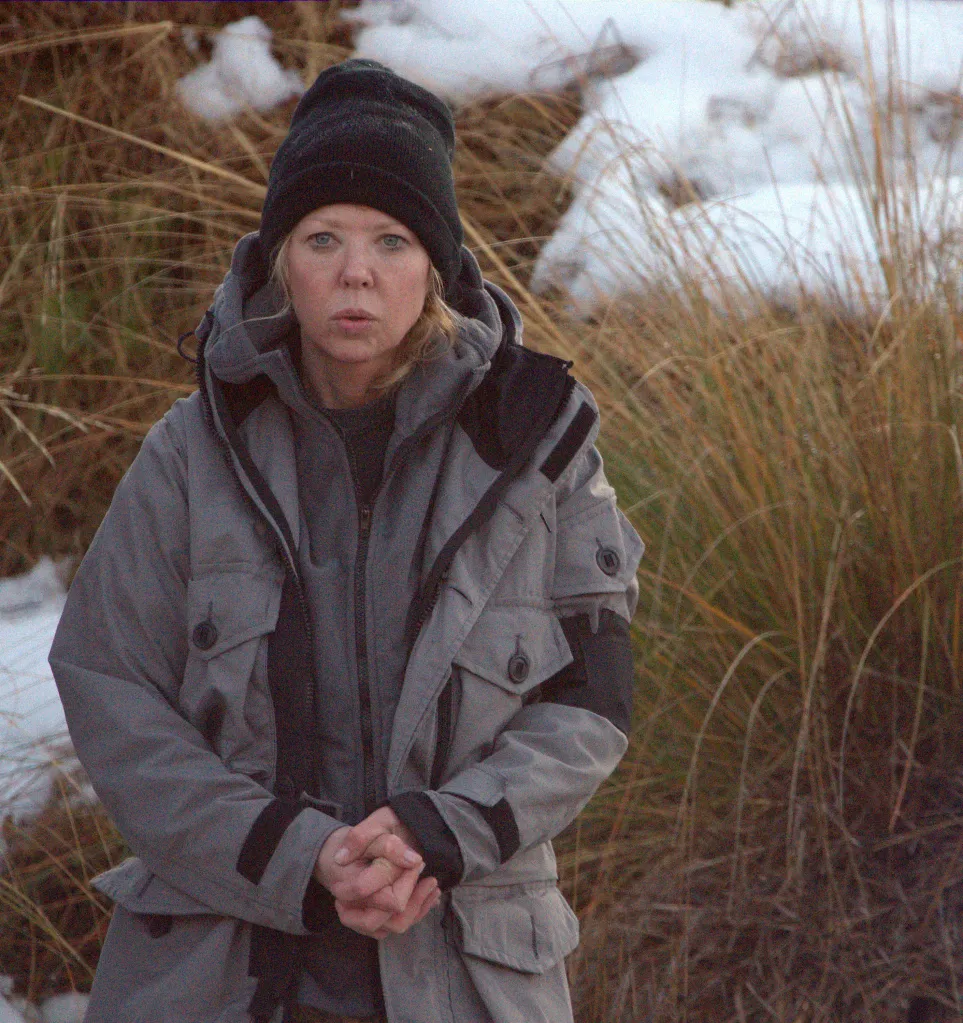 Tara Reid in a gray winter coat and black knit hat, with dry grass and snow in the background.