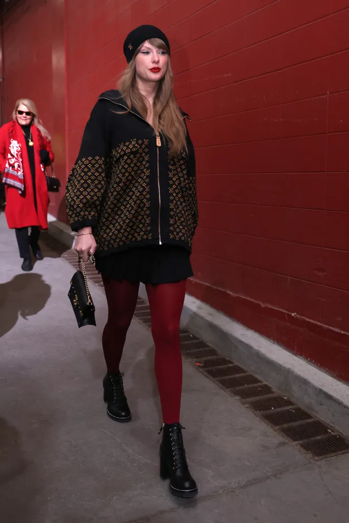 Taylor Swift wearing a black beanie, black zip-up jacket with gold LV patterns, a black skirt, red tights, and black boots.